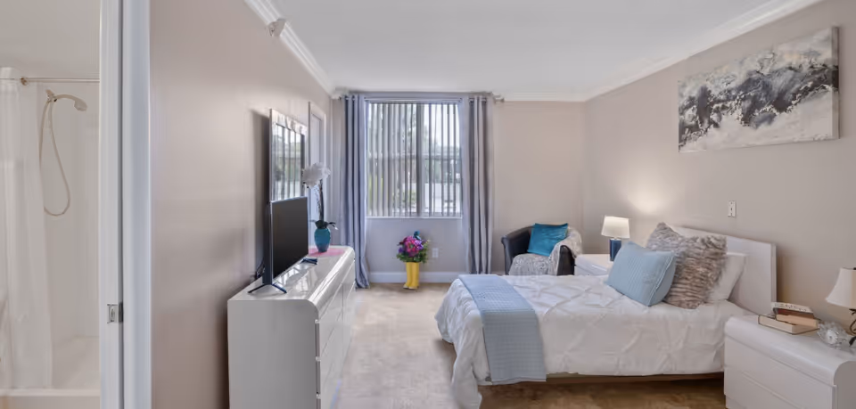 Bright furnished bedroom with a bed, nightstands and a TV on a dresser, an armchair by a window, and a bathroom doorway to the left.