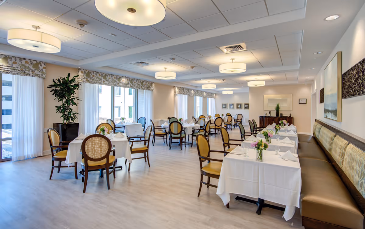 A bright and spacious dining room with multiple tables covered in white tablecloths, each set with napkins, glasses, and small flower arrangements. The room features large windows with sheer curtains and patterned valances, wooden chairs with cushioned seats, and a long cushioned bench along one wall. Modern circular ceiling lights illuminate the space, and there are framed pictures and artwork on the walls.