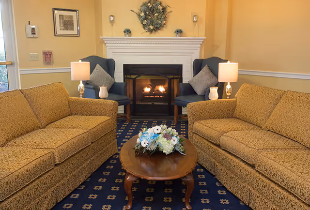 Cozy sitting area with two patterned sofas facing a lit fireplace, flanked by armchairs, lamps, and a coffee table with a floral arrangement.