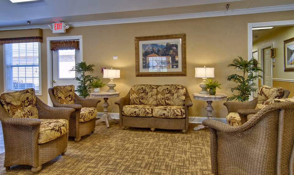 A cozy seating area in a senior living facility with wicker furniture including a loveseat and four armchairs, all with floral cushions. Two side tables with lamps and potted plants are placed beside the loveseat. A framed painting hangs on the beige wall behind the loveseat. There is a window and a door with an exit sign above it on the left side, and a hallway visible on the right.