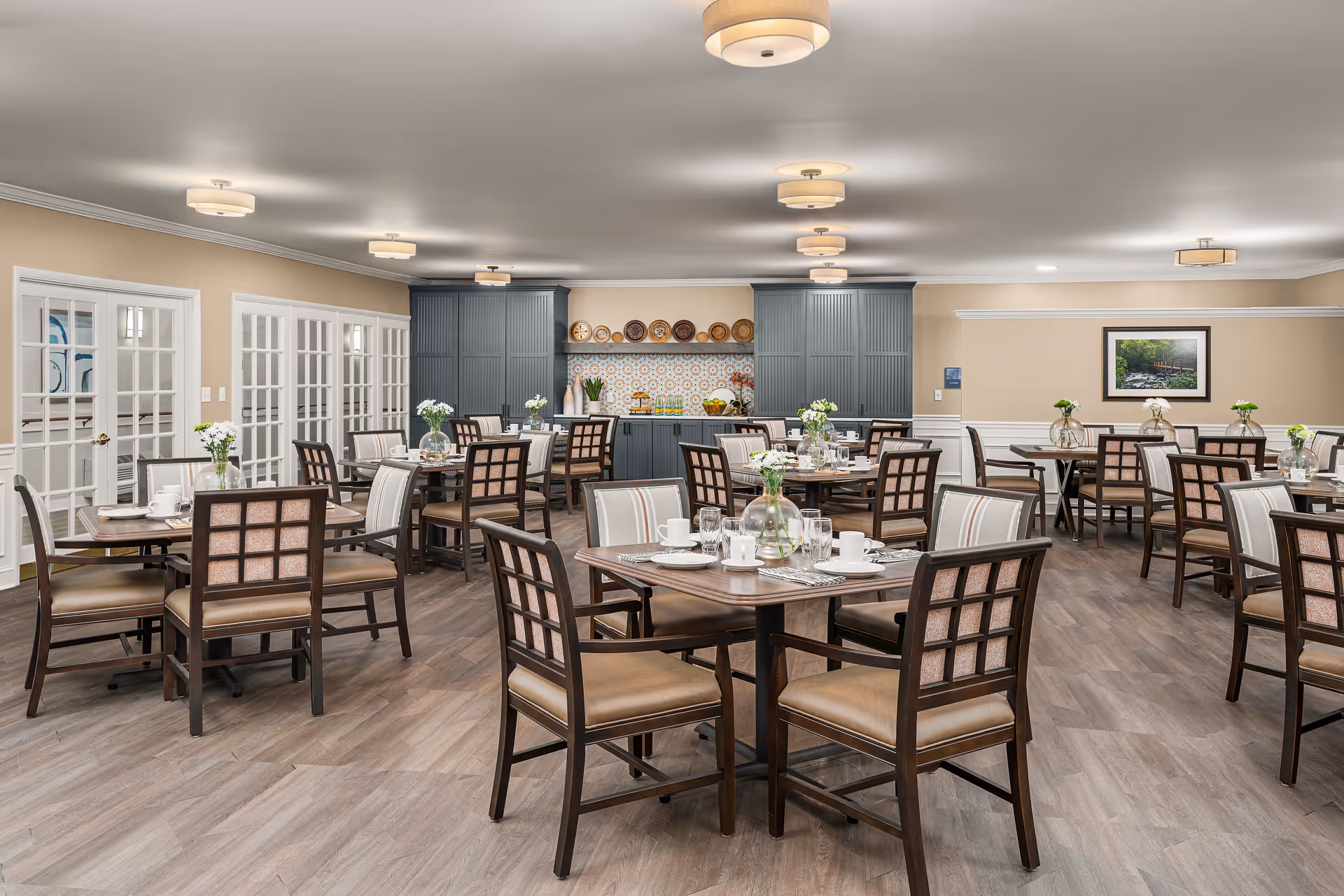 A spacious dining room with multiple wooden tables and chairs arranged neatly. Each table is set with white plates, cups, glasses, and silverware, and decorated with small vases holding white flowers. The room features light-colored walls, wood flooring, and ceiling lights. At the back, there are blue cabinets with decorative plates and a colorful tiled backsplash. French doors line one side of the room, and a framed picture hangs on the far wall.