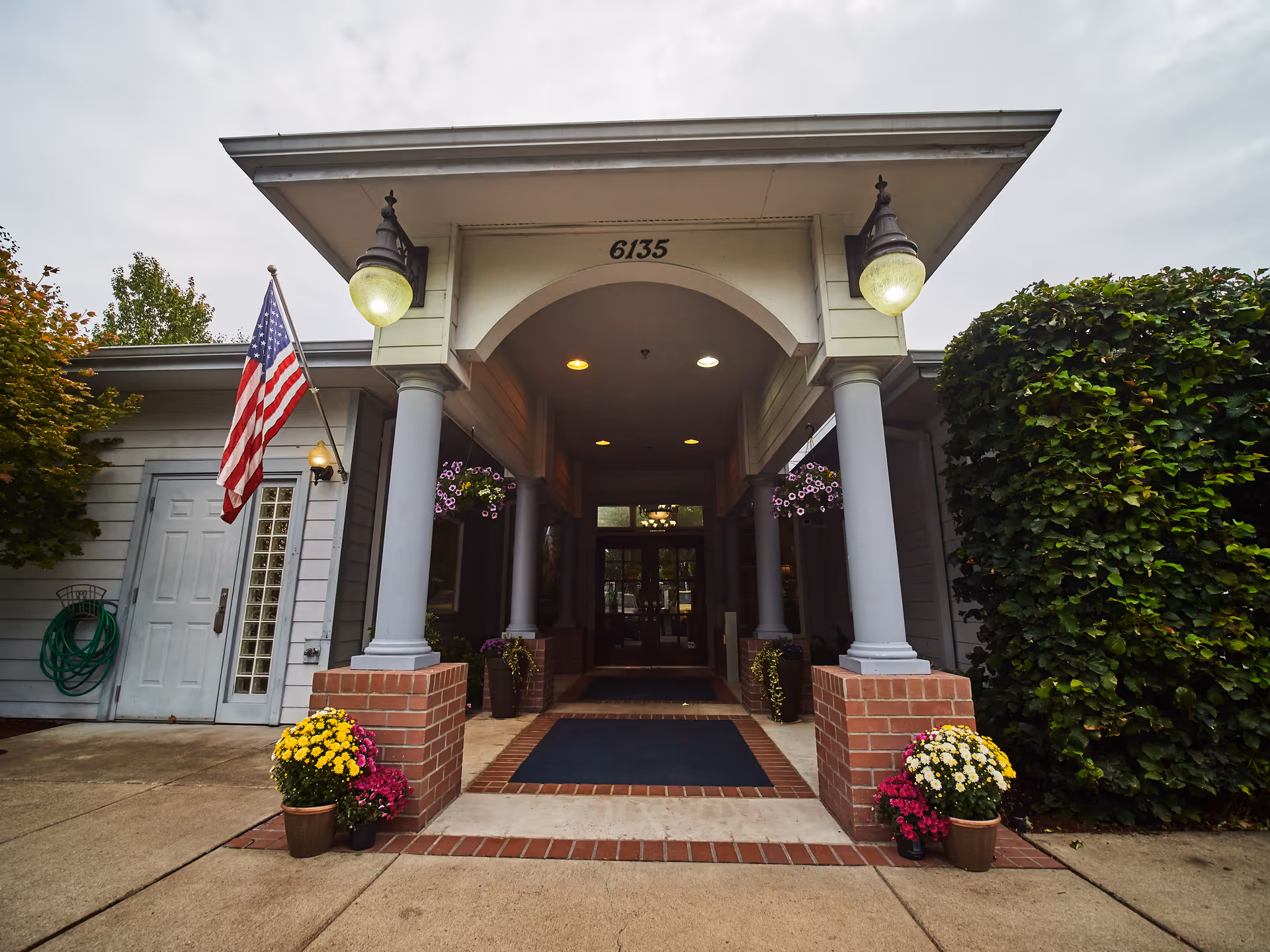 Entrance of a building with the number 6135 above the doorway, featuring a covered porch supported by four columns with brick bases. Two hanging flower pots and potted flowers on the ground flank the entrance. An American flag is mounted on the left side near a door with a glass block window. The sky is overcast.