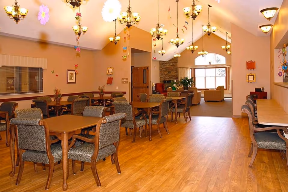 Spacious senior facility dining room with multiple wooden tables and upholstered chairs and a lounge area with sofas by a large window.