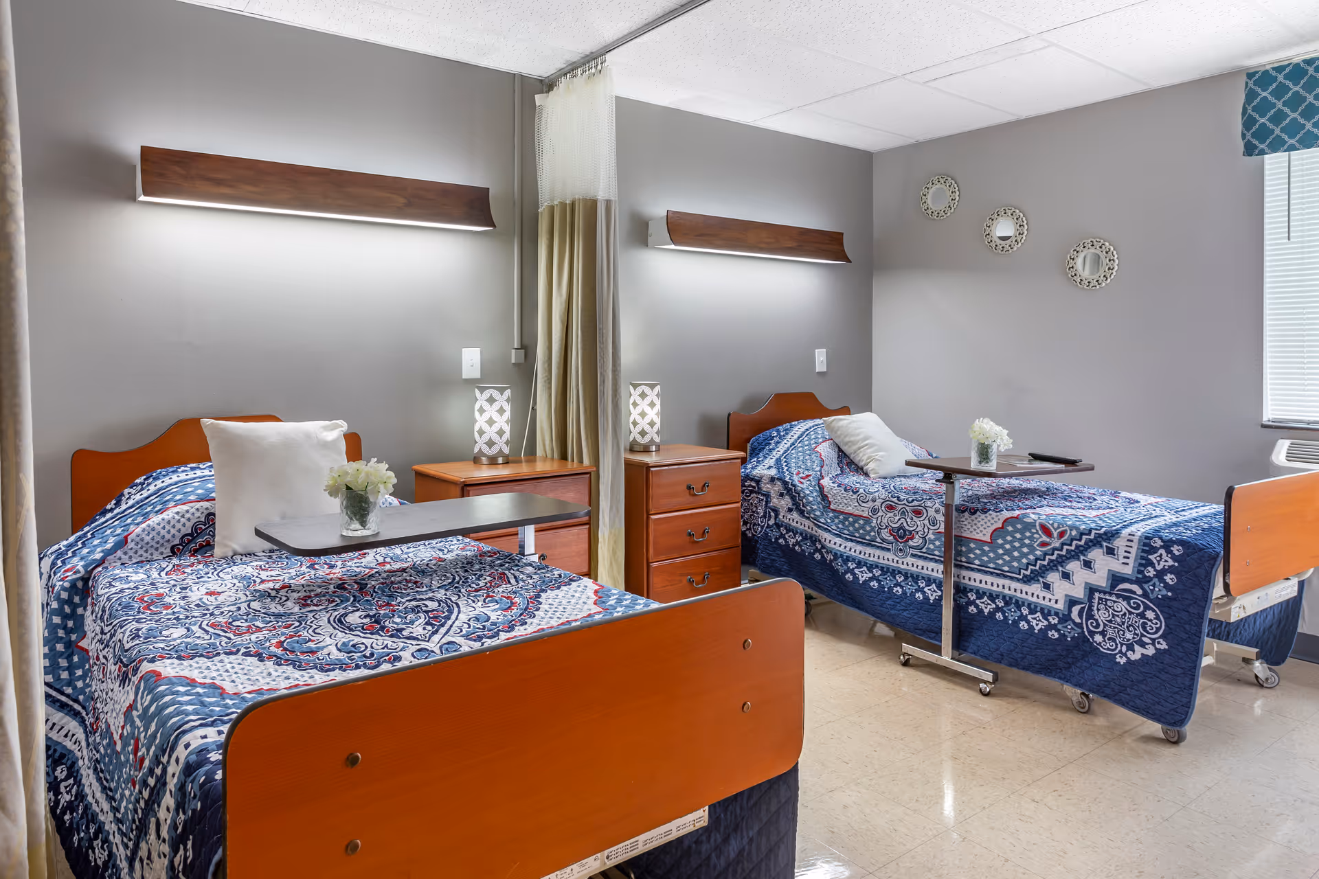 A shared bedroom in a senior living facility with two hospital-style beds, each covered with blue patterned quilts and white pillows. Between the beds is a wooden nightstand with a lamp. Each bed has an overbed table with a small vase of white flowers. The room has gray walls, three decorative round mirrors on one wall, and a window with blinds and a blue valance.