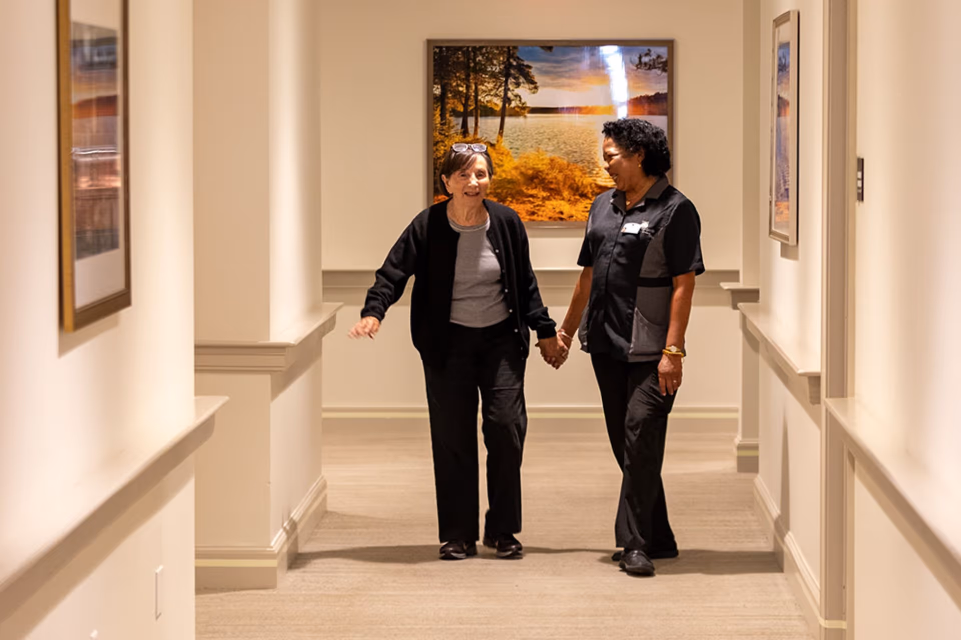 Two women walking hand in hand down a well-lit hallway with framed pictures on the walls. One woman appears to be a resident and the other a staff member, both smiling and engaged in conversation.