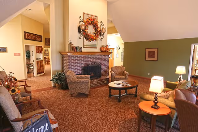 A cozy living room area in a senior living facility featuring a brick fireplace decorated with a fall-themed wreath and candles. The room has patterned carpet, several upholstered armchairs, a round wooden coffee table with magazines, and side tables with lamps. A sign reading 'Coffee Shop' is visible in the background through an open doorway.