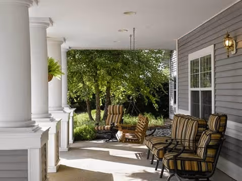 A covered porch area with white columns and cushioned outdoor chairs arranged along the side of a gray house. Trees and greenery are visible in the background, creating a peaceful outdoor setting.