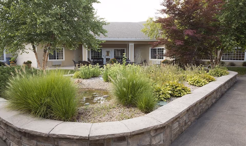 A landscaped garden area with various green plants and shrubs surrounded by a low stone wall, in front of a single-story building with a covered patio and outdoor seating.