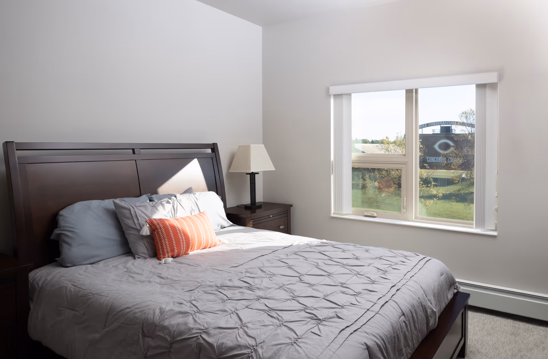 A bedroom with a neatly made bed featuring gray bedding and a decorative orange pillow. There is a dark wooden headboard and a matching nightstand with a lamp. A window with vertical blinds lets in natural light and shows a view of a green outdoor area with a sign that reads 'Concordia College'.