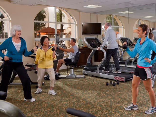 A group of older adults exercising in a fitness room with large windows. Two women are lifting dumbbells, a man is using a seated exercise machine, another man is walking on a treadmill, and a younger woman in athletic wear appears to be instructing or encouraging them. The room has carpeted floors and mirrors on the walls.