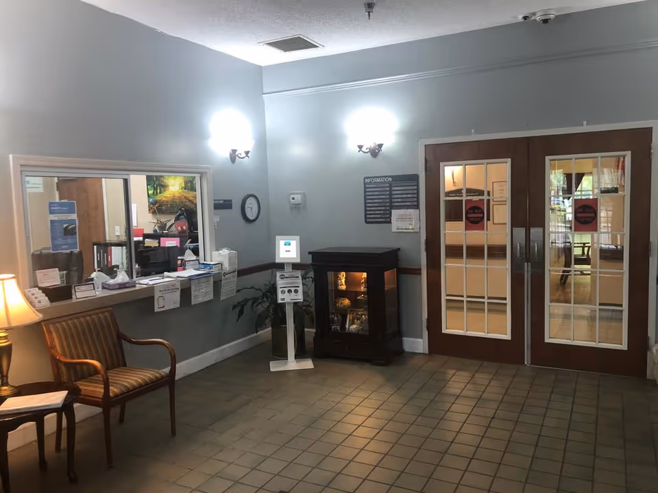 Interior view of a facility reception area with a wooden chair and small table with a lamp on the left, a reception window with papers and office supplies behind it, a clock on the wall, a small display cabinet with decorative items, and double wooden doors with glass panes and 'Do Not Enter' signs.