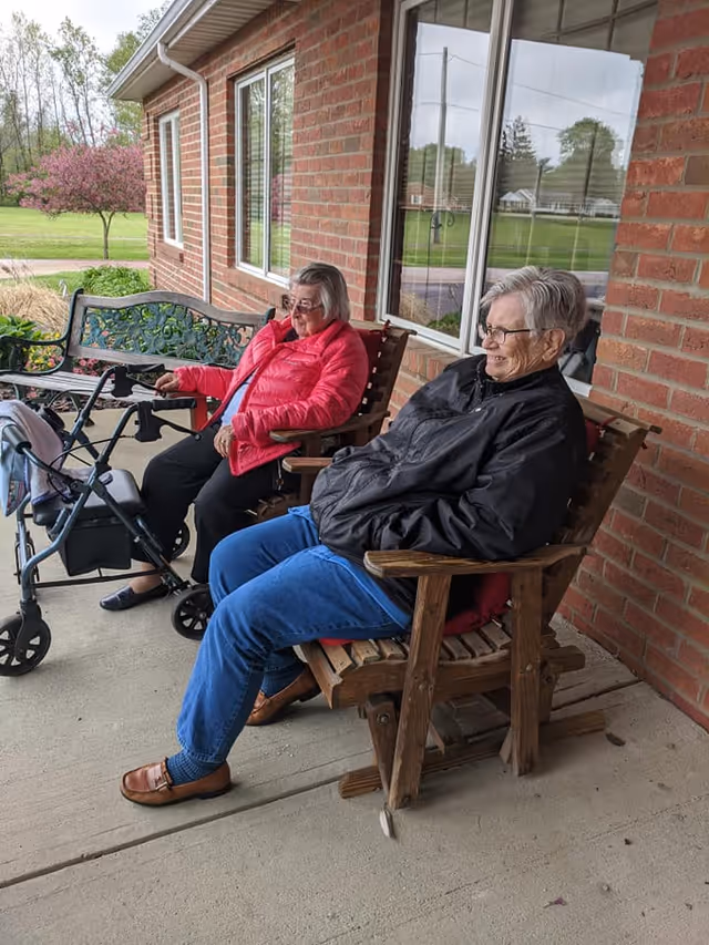 Two elderly people sit on wooden chairs on a covered brick porch with a walker beside them and a bench nearby.