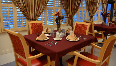 A dining area with a table covered in a burgundy tablecloth set for four people with beige napkins, white plates, cups, and silverware. The chairs have wooden frames with red patterned cushions. Behind the table are large windows with white shutters and striped curtains. A vase with flowers is placed in the center of the table.
