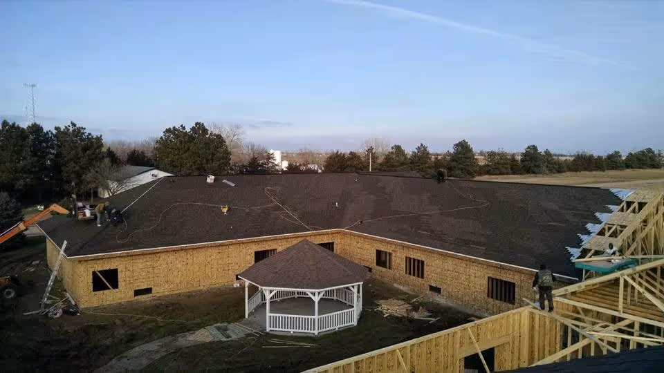 A large building under construction with workers on the roof and wooden framing visible. The building has a dark shingled roof and a white gazebo in the courtyard area. Surrounding the building are trees and open land under a clear sky.