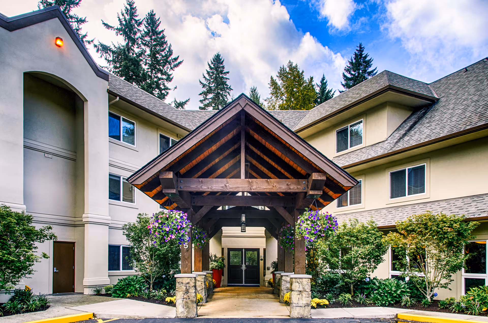 Front entrance of a multi-story senior living building with a wooden porte-cochere, hanging flower baskets, and landscaped shrubs.