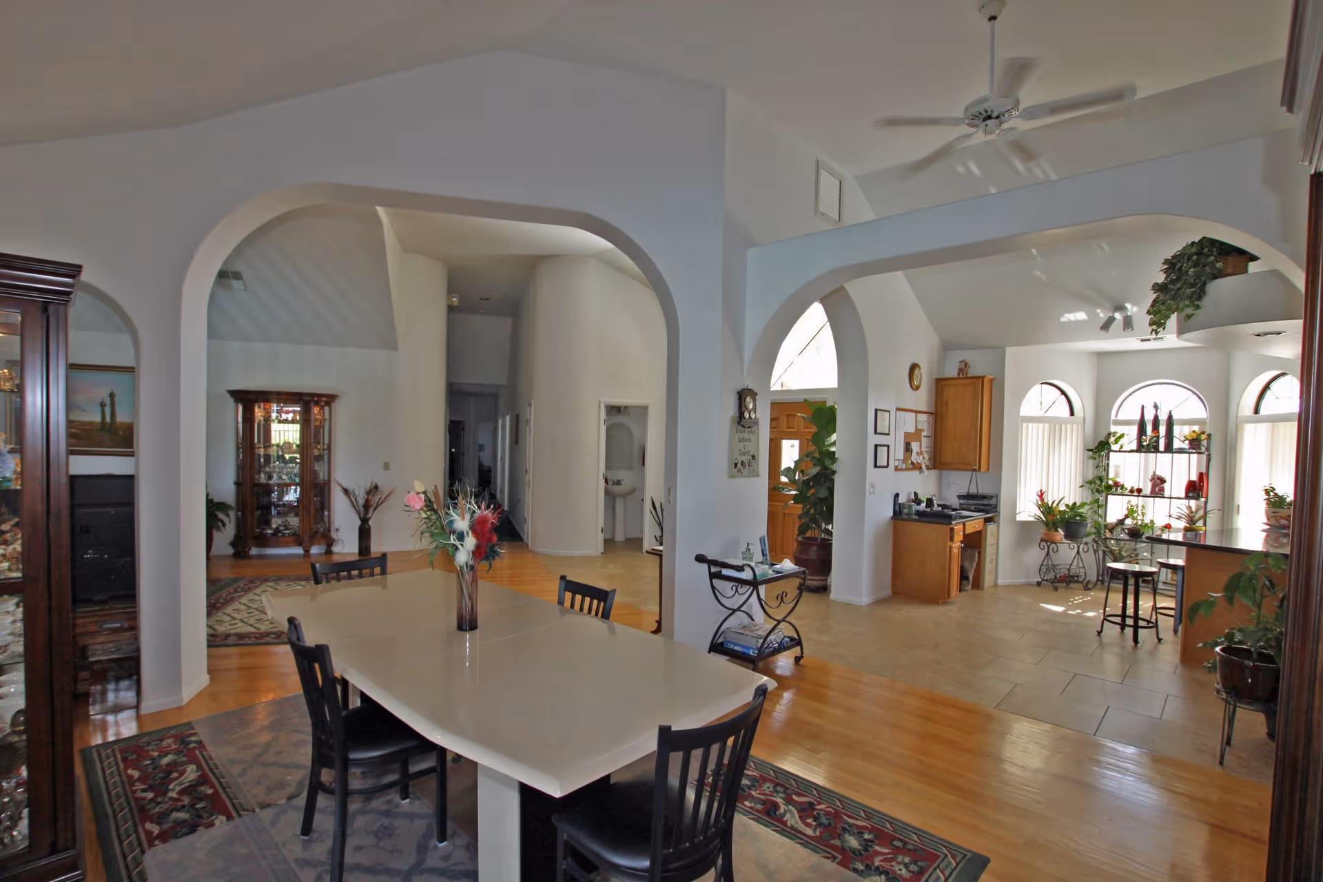 Interior view of a senior living facility showing a dining area with a rectangular table and black chairs on a patterned rug. The room has arched doorways leading to other areas including a kitchen with wooden cabinets and a tiled floor. There are plants and decorative items placed around the space, and a ceiling fan is visible above.