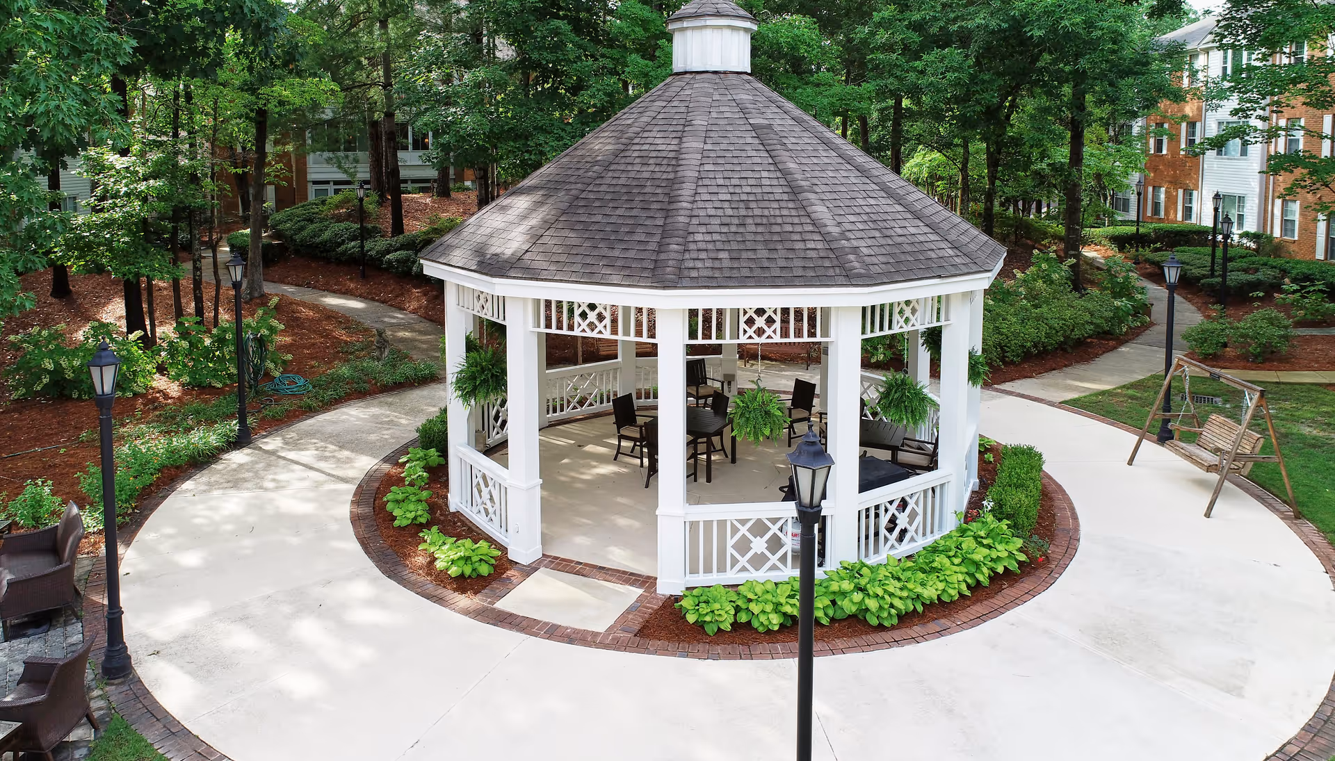 A white octagonal gazebo with a shingled roof situated in a landscaped outdoor area with paved walkways, green plants, and trees. The gazebo contains several chairs and hanging plants. Nearby, there is a wooden swing and outdoor seating. Surrounding the area are residential-style buildings partially visible through the trees.