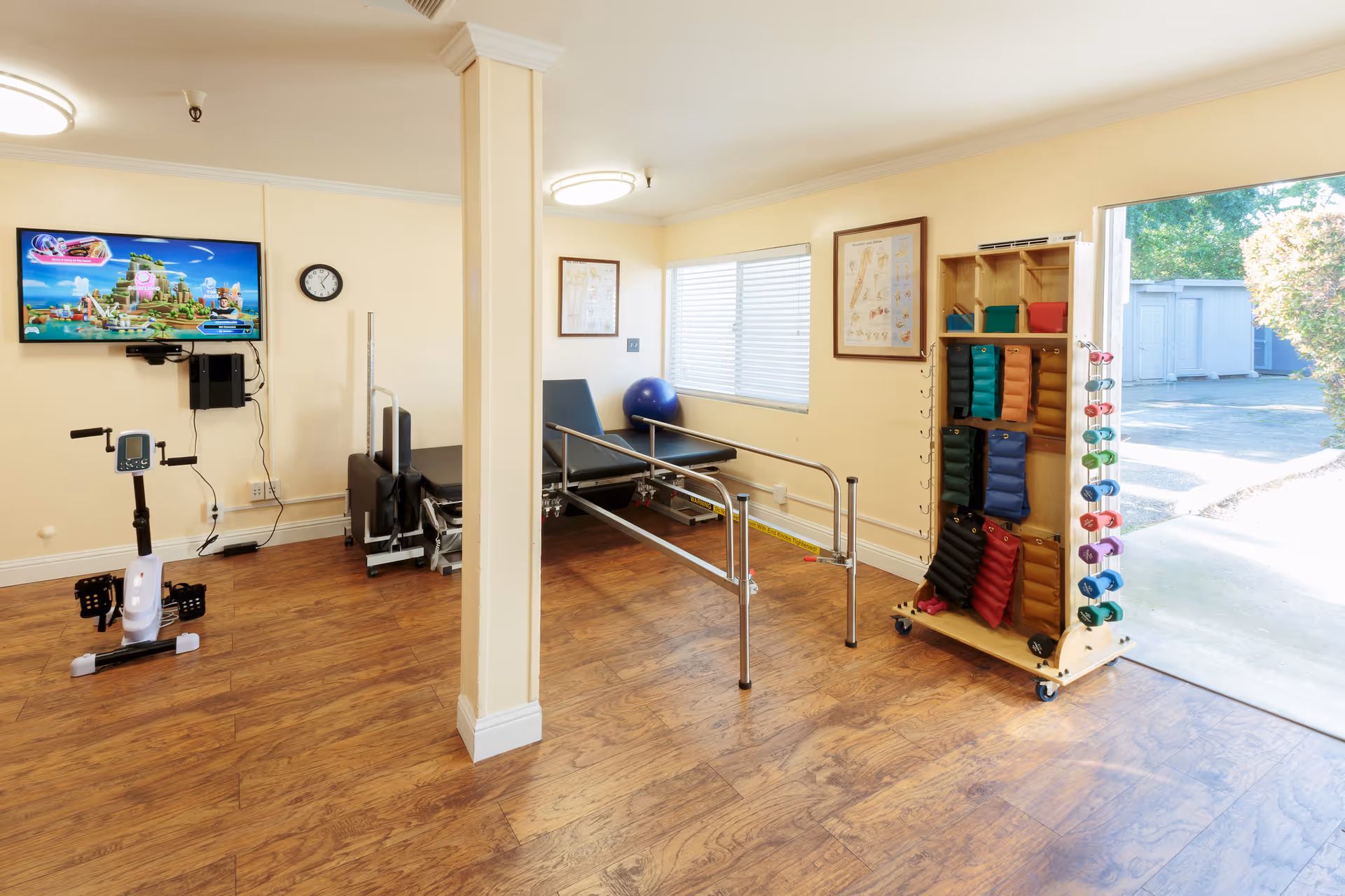 Bright therapy/exercise room with parallel bars, an exercise bike, a rack of weights and a wall-mounted TV, with a door open to the outside.