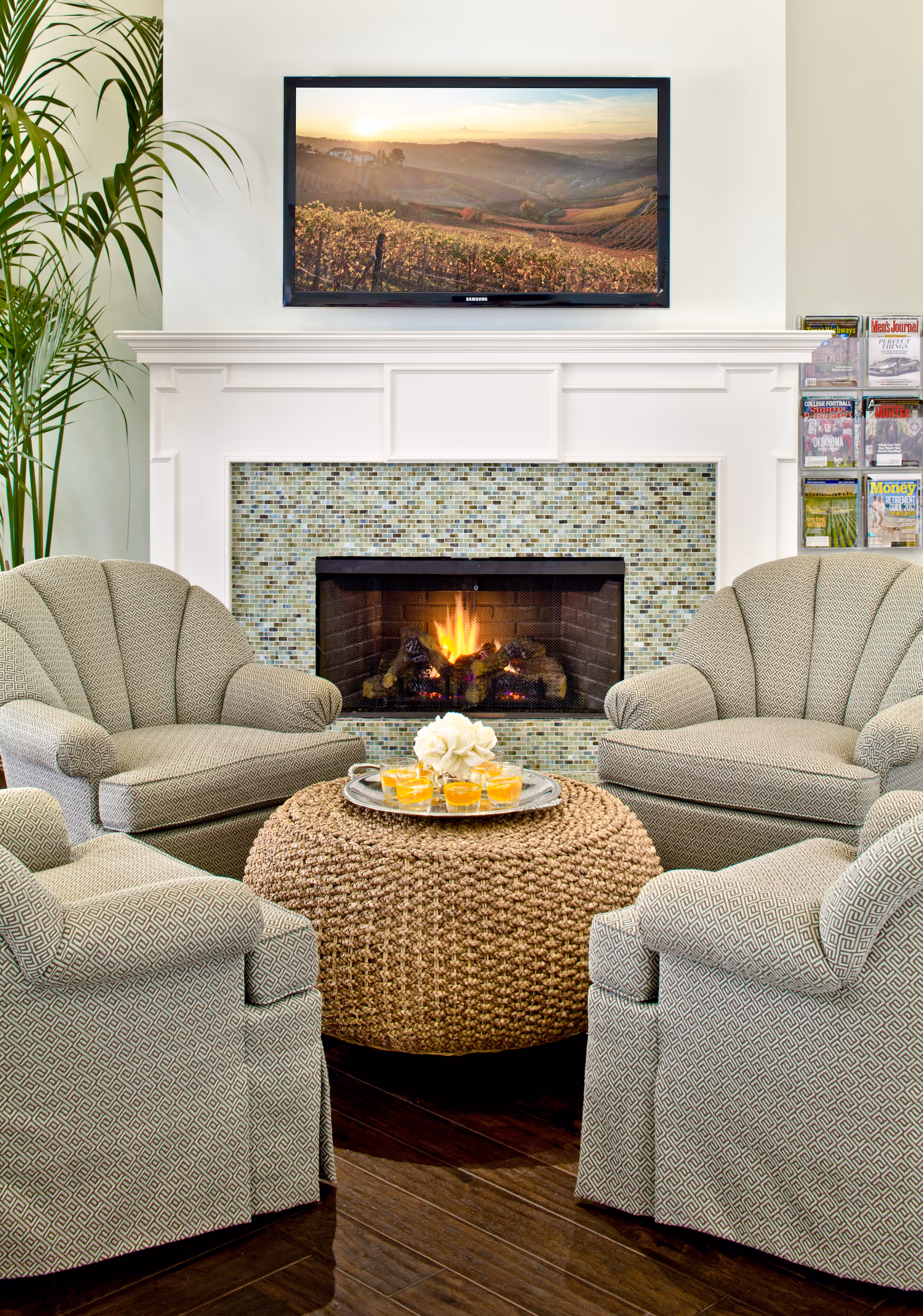 A cozy seating area with four patterned armchairs arranged around a round woven ottoman with a tray holding glasses and a flower. Behind the seating is a fireplace with a mosaic tile surround and a fire burning inside. Above the fireplace is a wall-mounted flat-screen TV displaying a scenic landscape of hills at sunset. To the right of the fireplace is a magazine rack filled with various magazines, and to the left is a tall green plant.