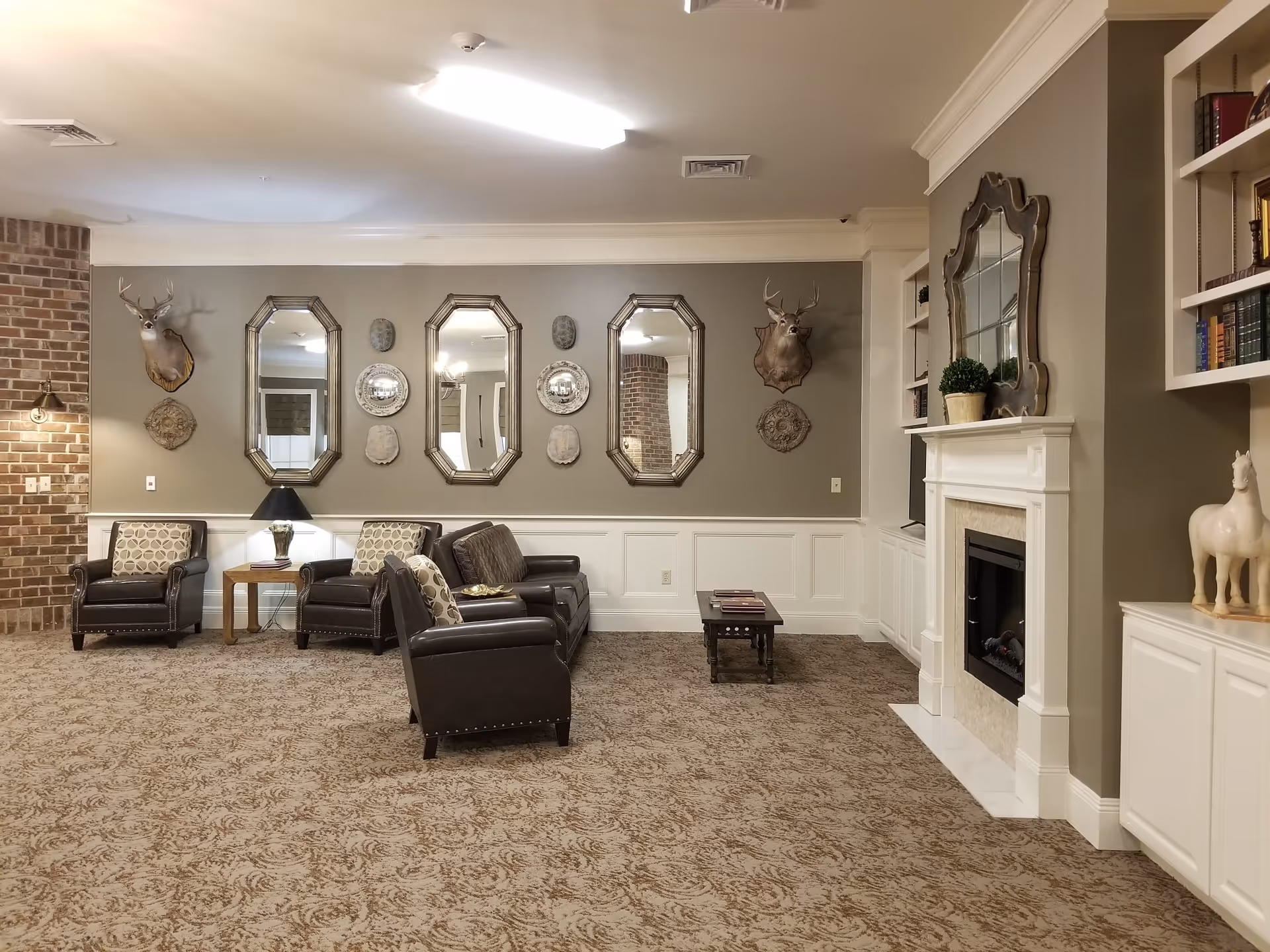 A cozy living room area with beige patterned carpet, three dark leather armchairs with patterned cushions, a small wooden side table with a lamp, a coffee table, and a white fireplace with a decorative mirror above it. The walls are decorated with three large mirrors, two mounted deer heads, and various decorative plates and plaques. Built-in white shelves with books and decorative items are on the right side.