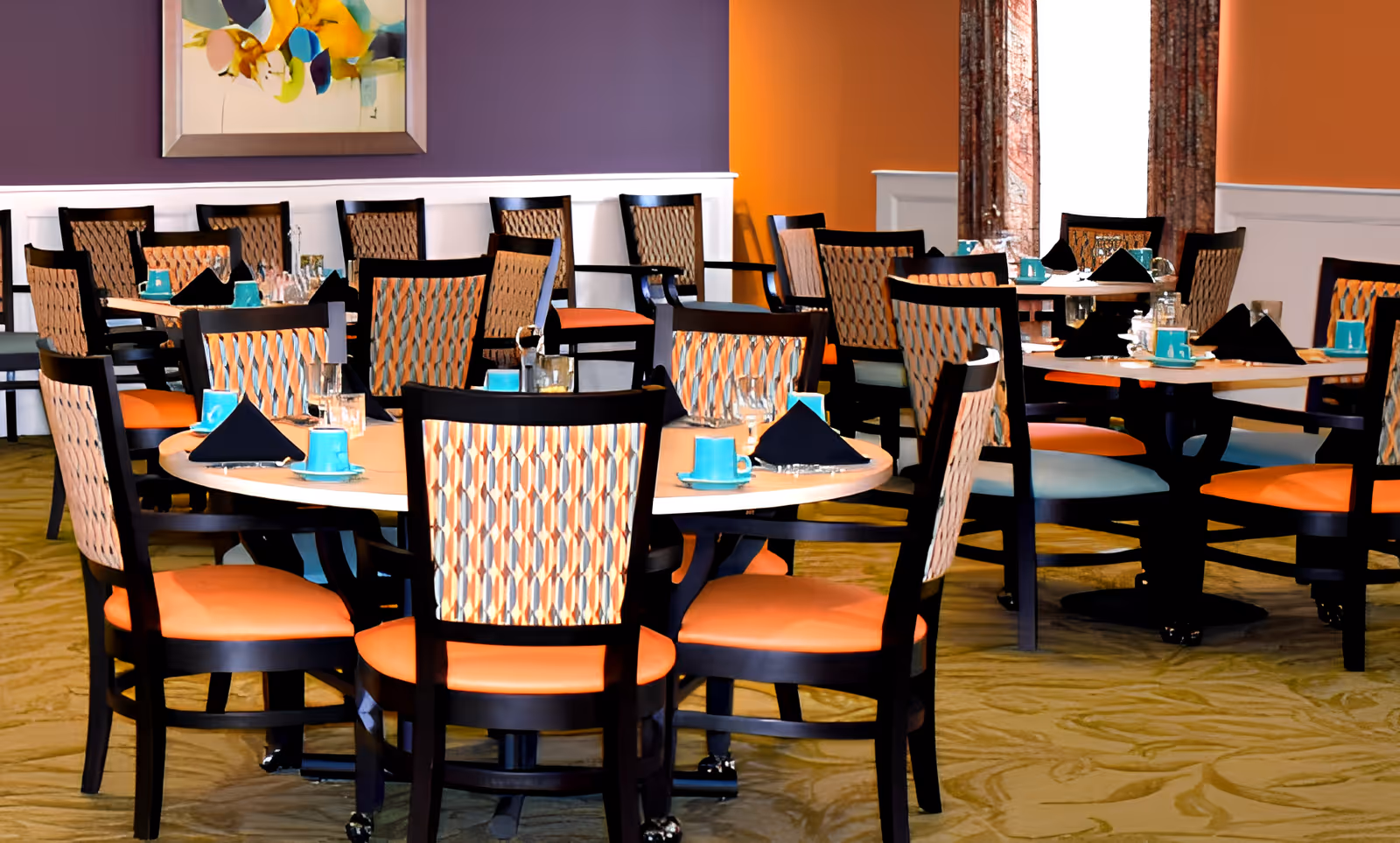 A dining room with multiple round tables set with turquoise cups, black napkins, and glassware. The chairs have patterned backs and orange seats. The walls are painted in purple and orange with white wainscoting, and there is a colorful abstract painting on the wall.