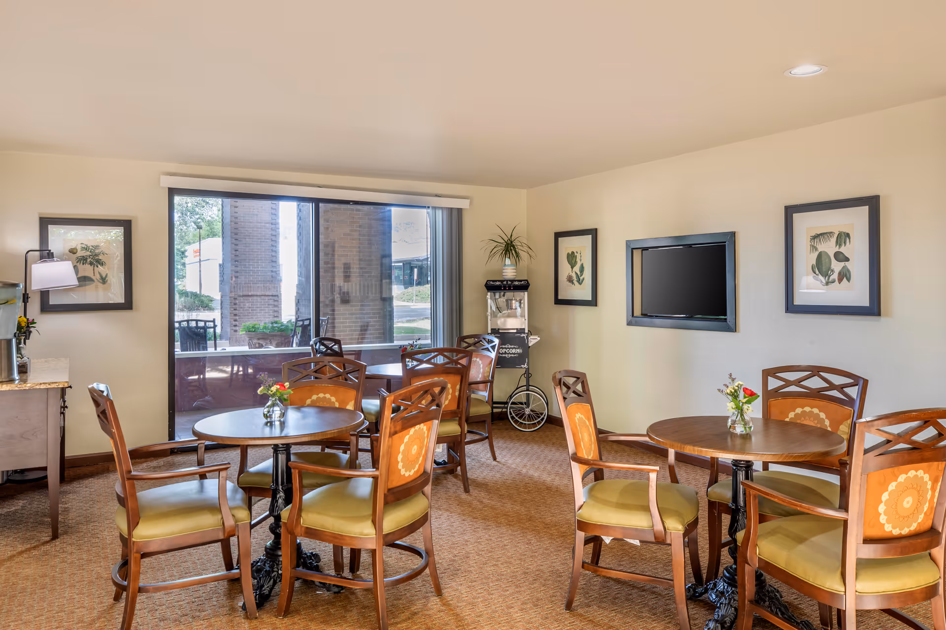 A cozy common area with round wooden tables and cushioned chairs arranged around them. There is a large window with a view of the outside, framed botanical artwork on the walls, a mounted flat-screen TV, and a popcorn machine in the corner. The room has warm lighting and a carpeted floor.