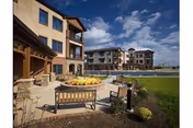 Outdoor seating area with wooden benches and a table decorated with pumpkins, situated in front of a multi-story senior living facility building under a partly cloudy sky.