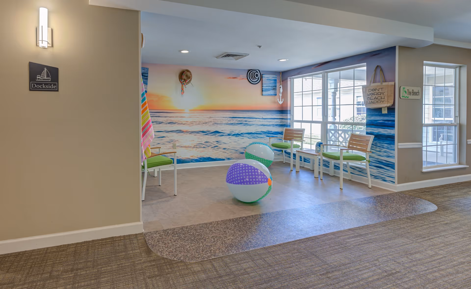 Indoor seating area with a beach theme, featuring a large mural of a sunset over the ocean on the wall. There are two beach balls on the floor, four chairs with green cushions, and two small white tables. Signs on the walls read 'Dockside' and 'The Beach,' and a tote bag hanging on the wall says 'Don't Worry Beachy Daddy.' Large windows let in natural light.