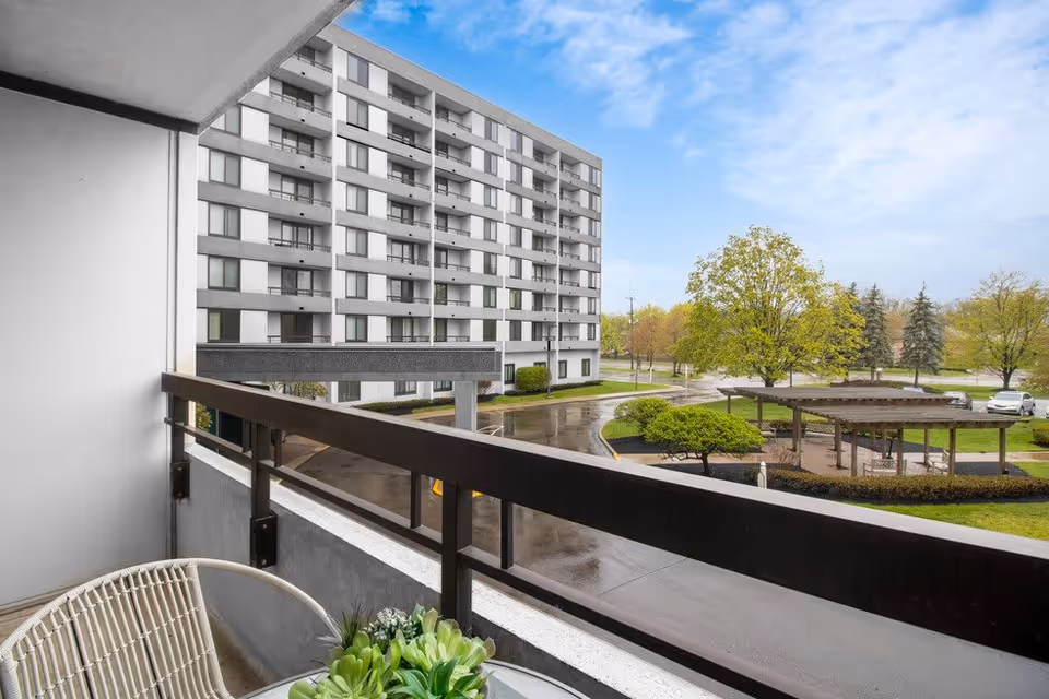 View from a balcony showing a multi-story residential building, a curved driveway, green trees, and a wooden pergola structure in a landscaped outdoor area under a partly cloudy sky.