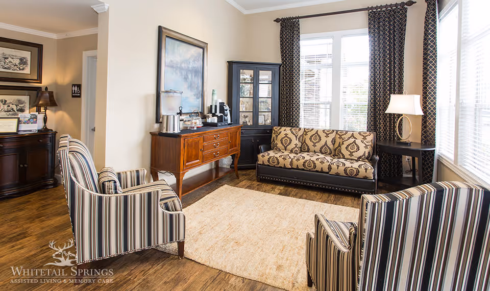 Sunlit sitting room with a patterned sofa, two striped armchairs, a wooden sideboard and large windows.