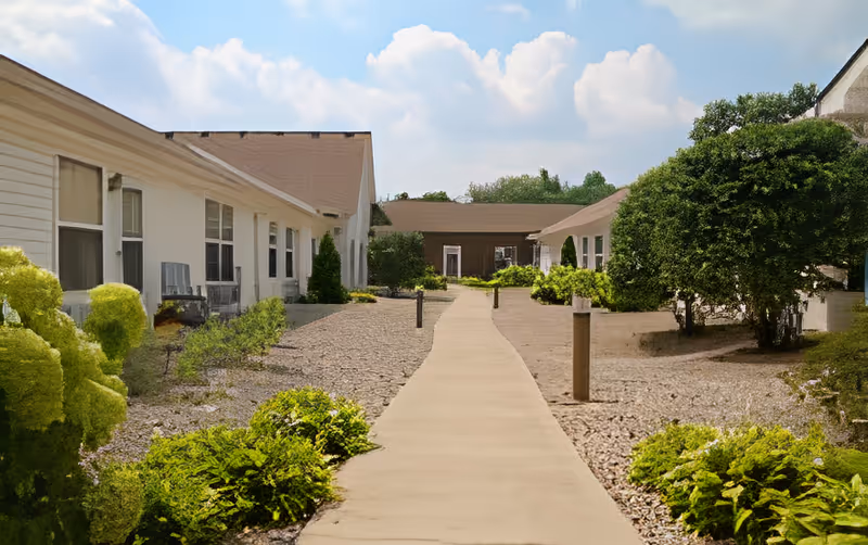 Outdoor walkway between two single-story buildings with white siding and brown roofs, surrounded by green bushes and small trees under a partly cloudy sky.