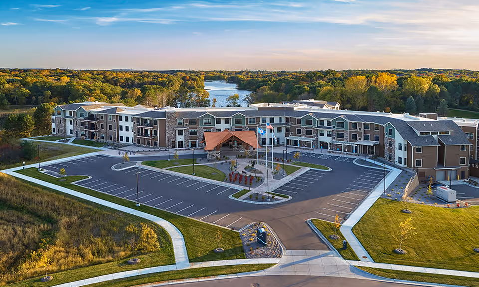 Aerial view of The Lakes at Stillwater senior living facility showing a large three-story building with a covered entrance, surrounded by a spacious parking lot and landscaped green areas. In the background, there is a river and dense trees with autumn foliage under a clear blue sky.