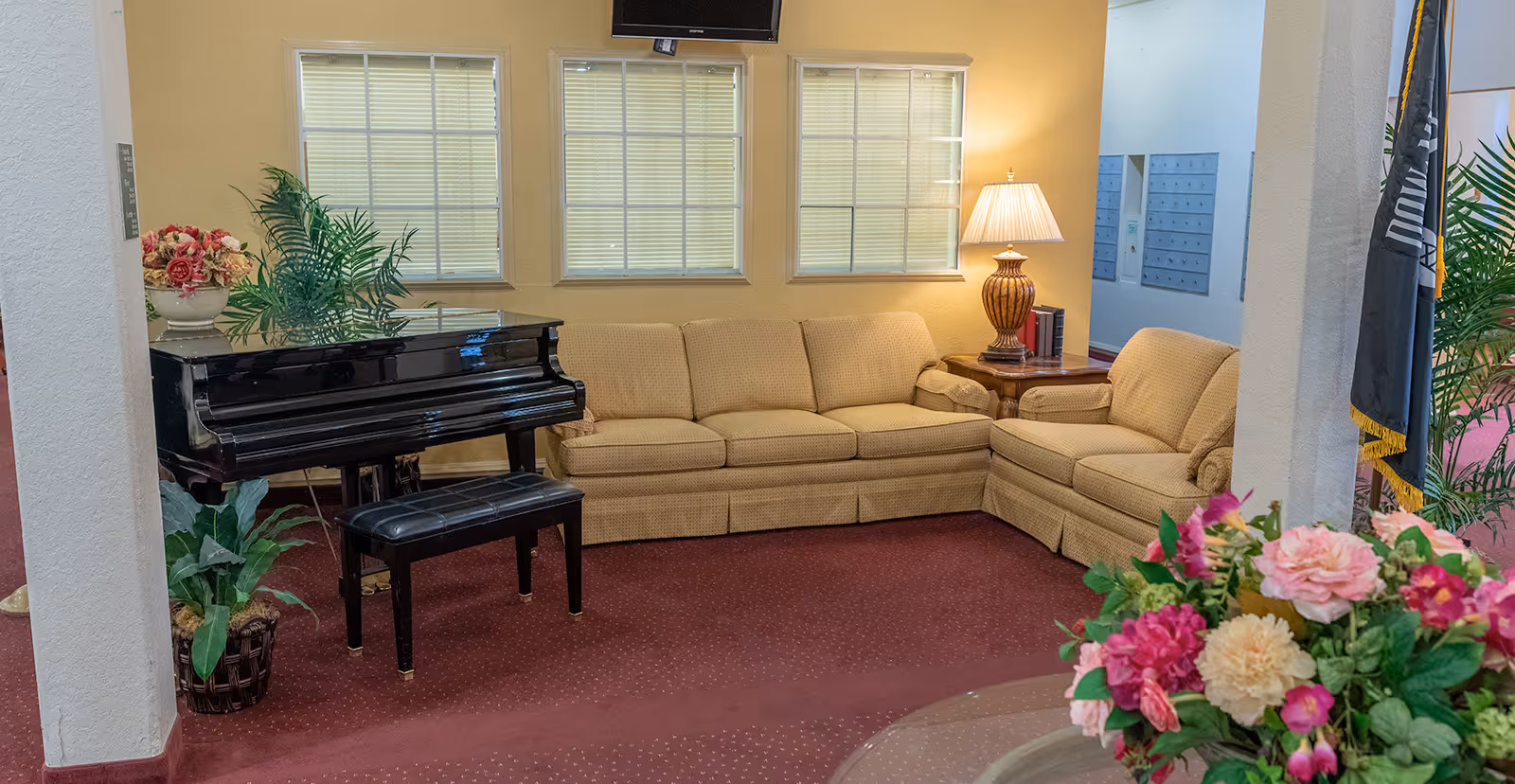 Carpeted common room with a beige sectional sofa, black grand piano, lamp, and floral arrangements.