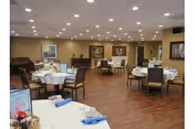 Spacious dining room with round tables set with white tablecloths and napkins, chairs, wood flooring, and recessed ceiling lights.