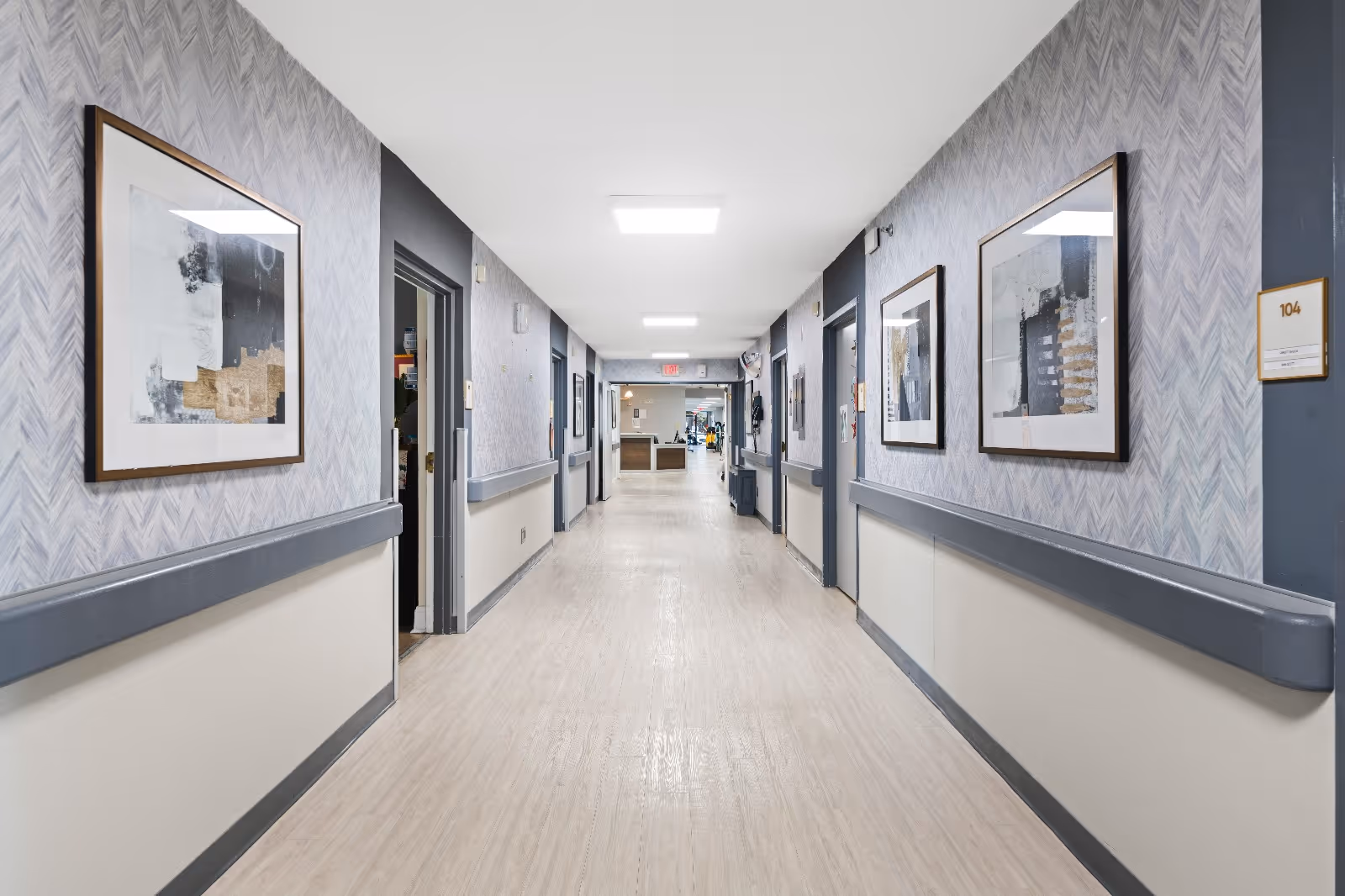 A clean, well-lit hallway in a senior living facility with light wood flooring, gray and white walls, and framed abstract artwork on both sides. Several doorways line the corridor, and there is a reception or common area visible at the far end.
