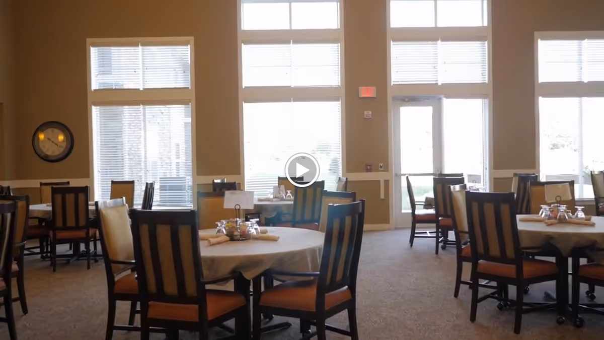 A dining room with multiple round tables covered with beige tablecloths, each surrounded by wooden chairs with cushioned seats. Large windows with blinds allow natural light to fill the room. A wall clock is visible on the left side.