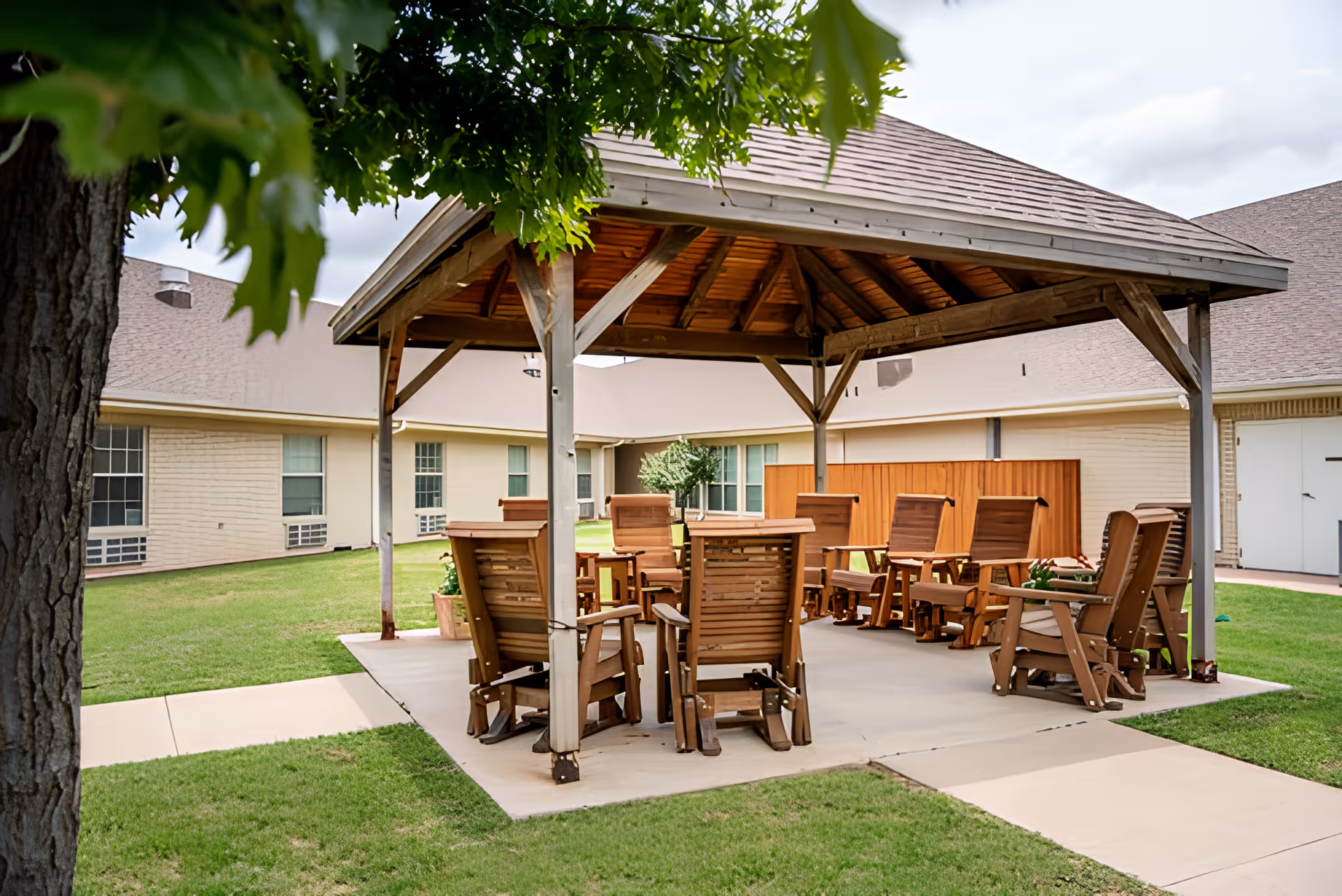 Wooden gazebo with chairs and tables on a concrete pad in a grassy courtyard beside a single-story building.