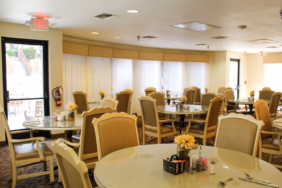 A bright dining room with multiple round tables and beige cushioned chairs. Each table has a small flower arrangement and condiments. Large windows with sheer curtains allow natural light to fill the room. There are exit doors visible on two sides of the room.