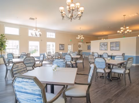 A bright and spacious dining room with multiple tables and chairs arranged neatly. The chairs have blue and white patterned upholstery. Large windows allow natural light to fill the room, and several chandeliers hang from the ceiling. The walls are decorated with framed artwork and painted in light colors with a blue accent.