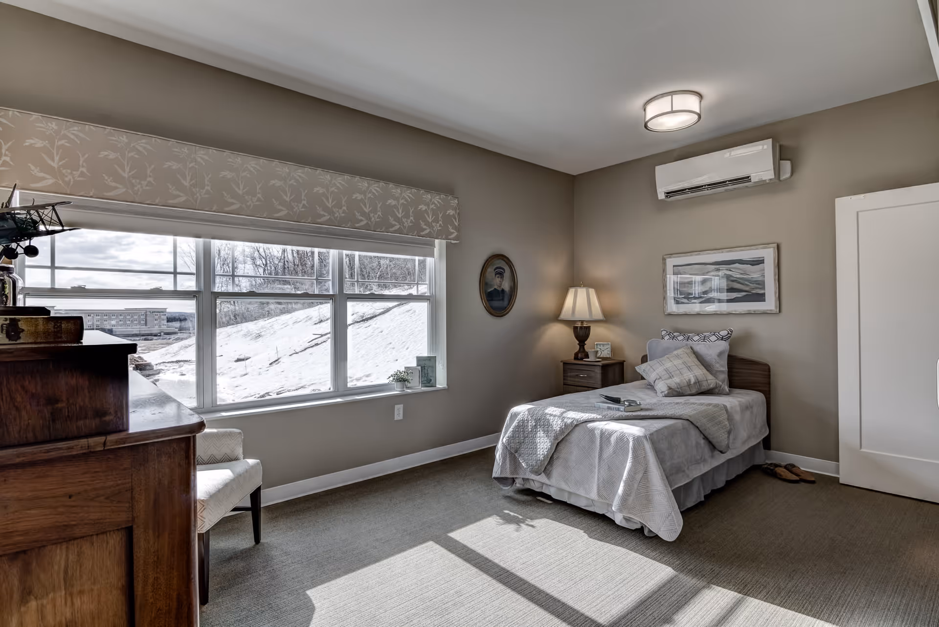A cozy bedroom with a single bed covered in light gray bedding and pillows. Next to the bed is a wooden nightstand with a lamp and small decor items. A large window with a floral valance lets in natural light, showing a snowy outdoor scene. There is a wooden dresser on the left side and a white chair near the window. The walls are painted beige and decorated with framed artwork and a portrait. A white door and a wall-mounted air conditioning unit are also visible.