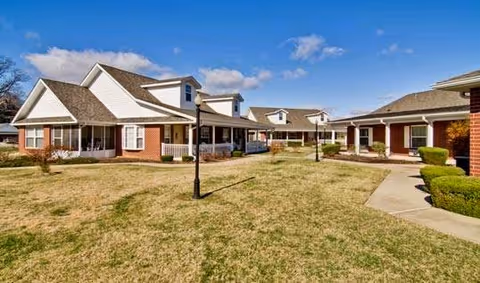 Exterior view of a senior living facility with multiple single-story buildings featuring brick and white siding, pitched roofs, and covered porches surrounding a grassy courtyard with a paved walkway and a lamp post under a blue sky with some clouds.