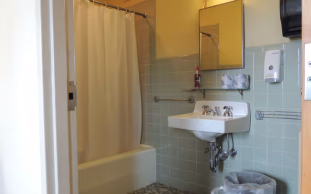 A bathroom with a white bathtub and shower curtain on the left, a wall-mounted white sink with chrome faucets, a mirror above the sink, a soap dispenser, a shelf with a bottle, and a trash can below the sink. The walls are tiled in light green and the floor has a patterned tile.