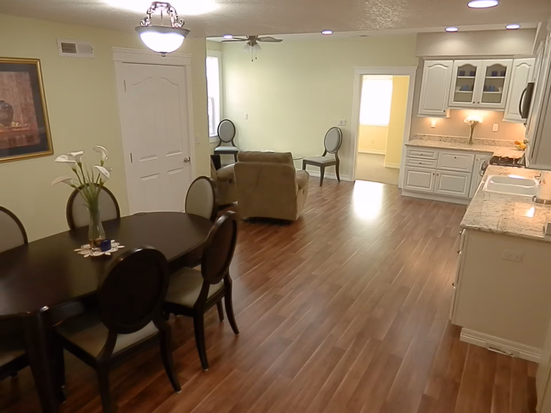 Interior view of a senior living facility showing a dining area with a dark wooden table and six chairs, a small sitting area with two chairs and an armchair, and a kitchen with white cabinets and granite countertops. The floor is wooden, and the walls are light-colored with a framed picture and a ceiling light fixture.