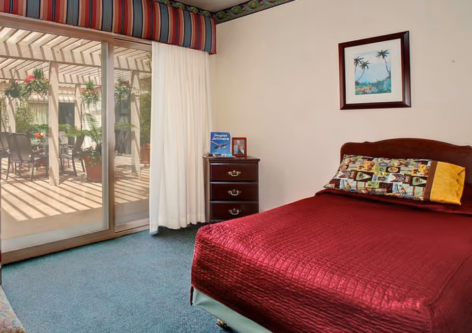 A bedroom with a burgundy-covered bed, a three-drawer nightstand, sliding glass doors opening to a pergola-covered patio, and framed art on the wall.