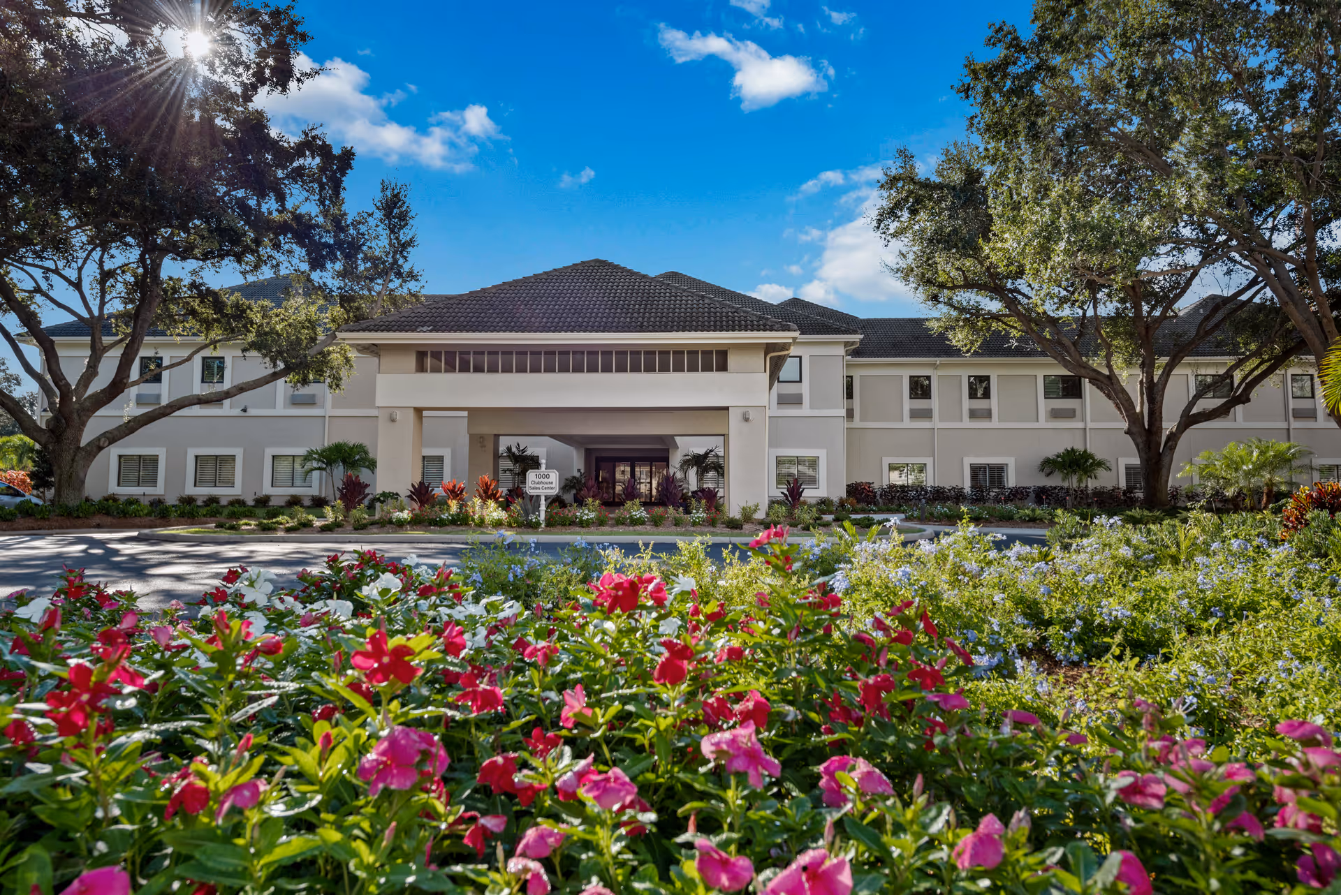 Front exterior view of Arbor Trace facility with a clear blue sky, sunlight filtering through trees, and colorful flowers in the foreground.