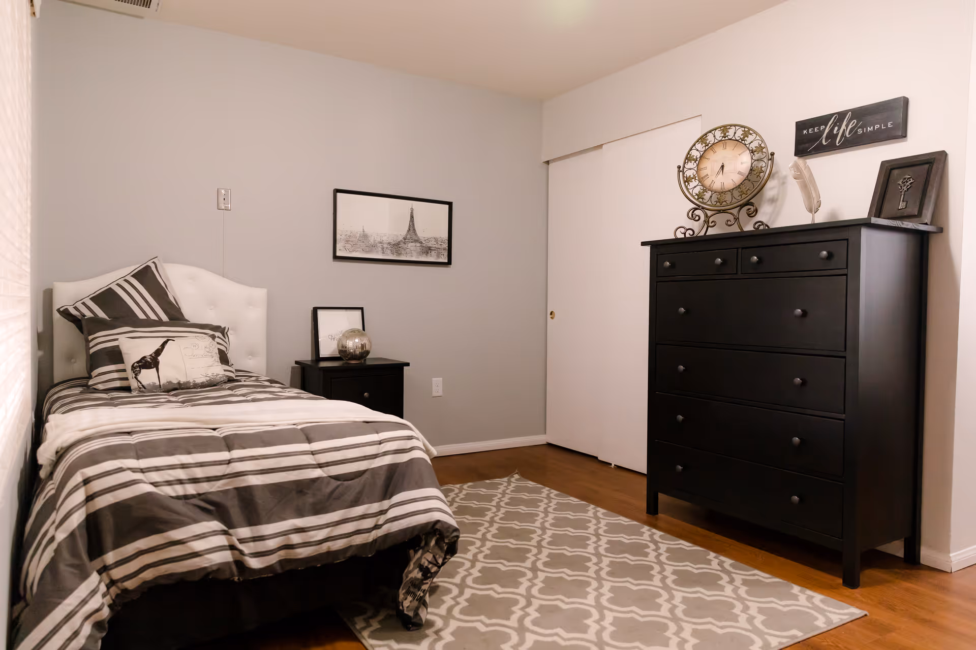 A neatly arranged bedroom with a single bed featuring striped bedding and decorative pillows, a black nightstand with a framed picture and a decorative globe, a black dresser with a vintage-style clock and framed art on top, a patterned area rug on a wooden floor, and light gray walls with a framed picture of the Eiffel Tower.