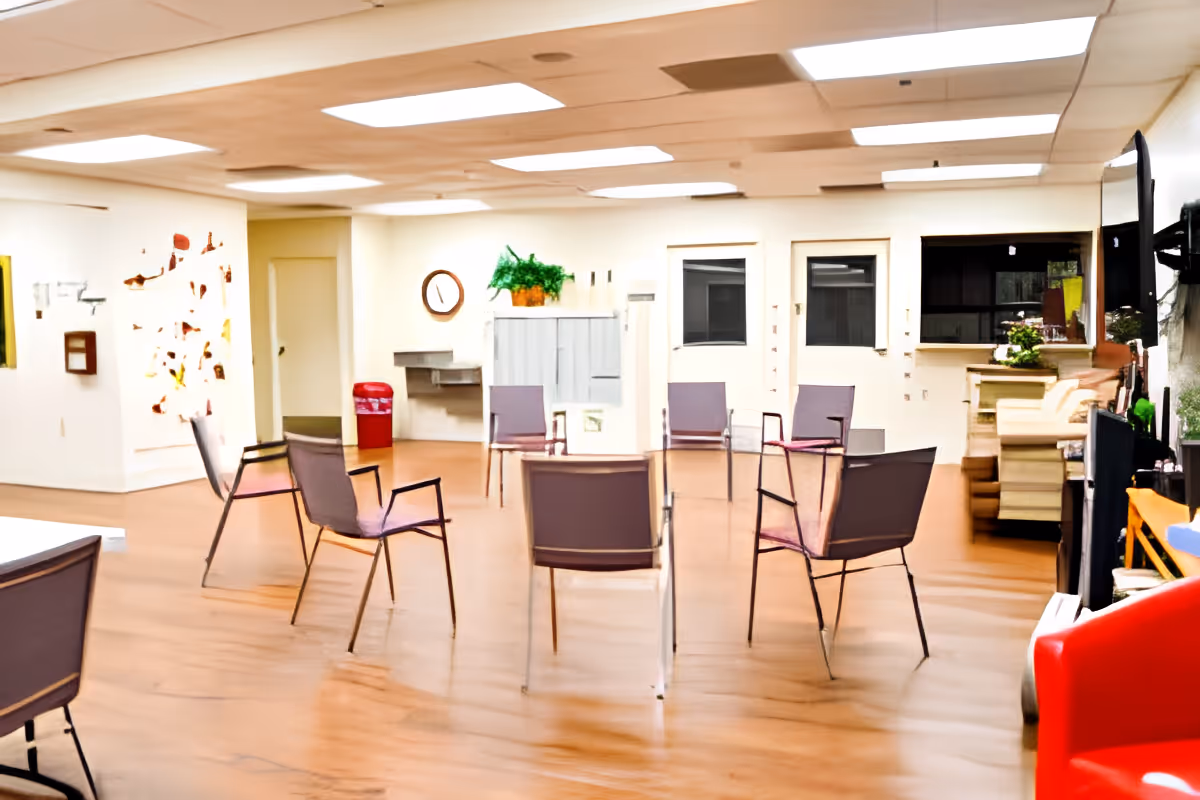 A spacious room with several chairs arranged in a circle on a wooden floor. The room has white walls, a clock on the wall, a red trash bin, a water fountain, and some plants. There is a large TV mounted on the right wall and some shelves with decorative items.