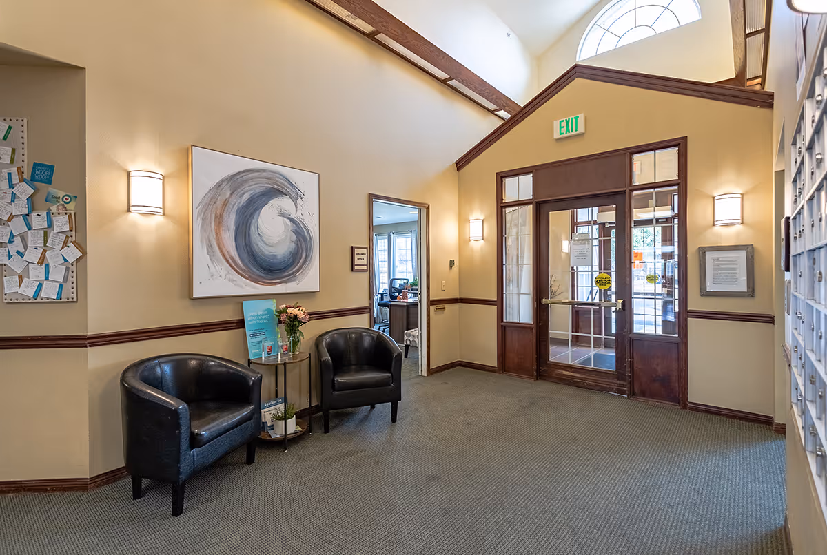 Interior view of a senior living facility lobby area with two black leather chairs, a small round table with flowers and informational brochures, a large abstract painting on the wall, a bulletin board with notes, and a glass door exit with an exit sign above it.