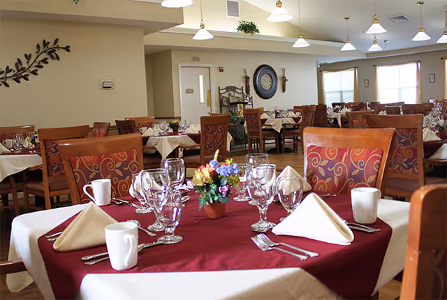A dining room in a senior living facility with tables set for a meal. Tables are covered with white and burgundy tablecloths, each set with folded napkins, glassware, coffee mugs, and silverware. Chairs have patterned cushions, and there is a small flower arrangement centerpiece on the table in the foreground. The room is well-lit with hanging lights and has windows letting in natural light.