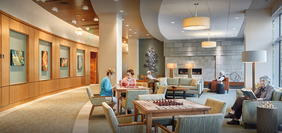 A spacious and well-lit senior living common area with several elderly residents engaged in different activities. Two women are sitting at a table playing a board game, while others are seated on sofas and chairs reading or relaxing near a modern fireplace. The room features contemporary decor with wooden paneling, artwork on the walls, and large windows allowing natural light to fill the space.